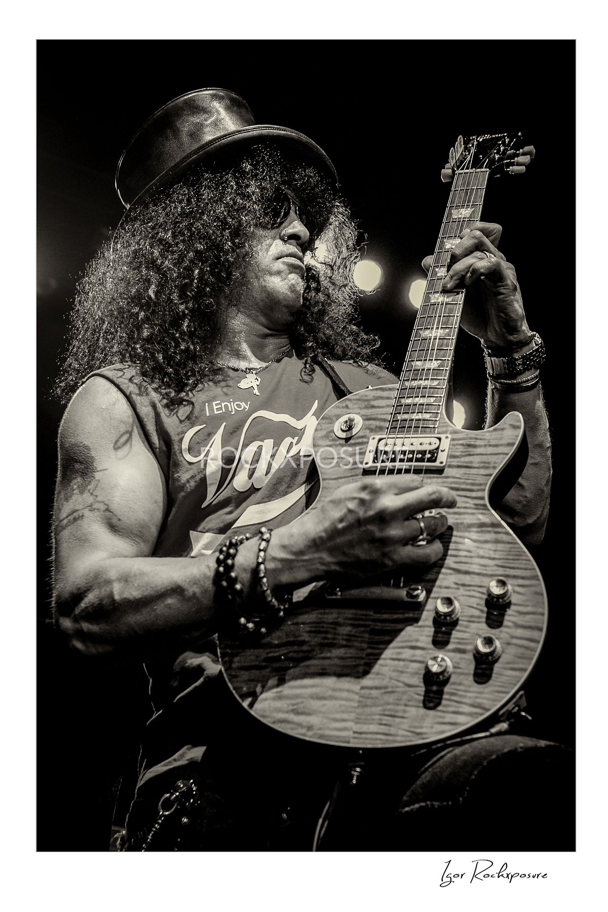 Vertical concert photography of Slash playing electric guitar in black and white photography wearing his signature top hat under dramatic stage lighting