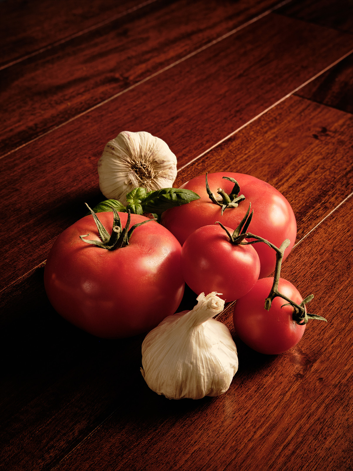 Several red tomatoes with garlic bulbs and basil leaves on mahogany wood.