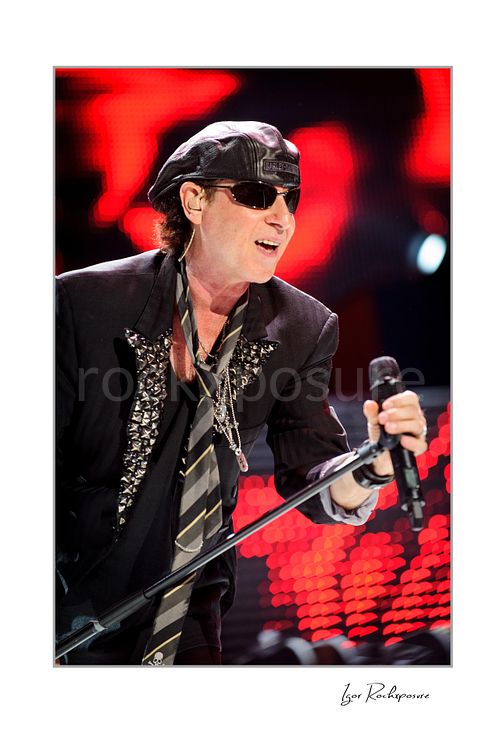 Vertical color image of Klaus Meine of Scorpions singing live in sunglasses and leather cap with microphone stand against red stage lights
