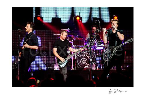 Horizontal color image of Nickelback performing onstage with Ryan Peake, Mike Kroeger, Chad Kroeger, and Daniel Adair under red concert lighting