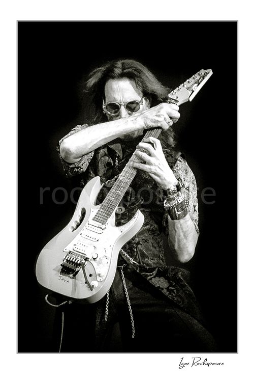 Vertical black and white image of Steve Vai playing guitar in dark sunglasses with a dramatic hand pose