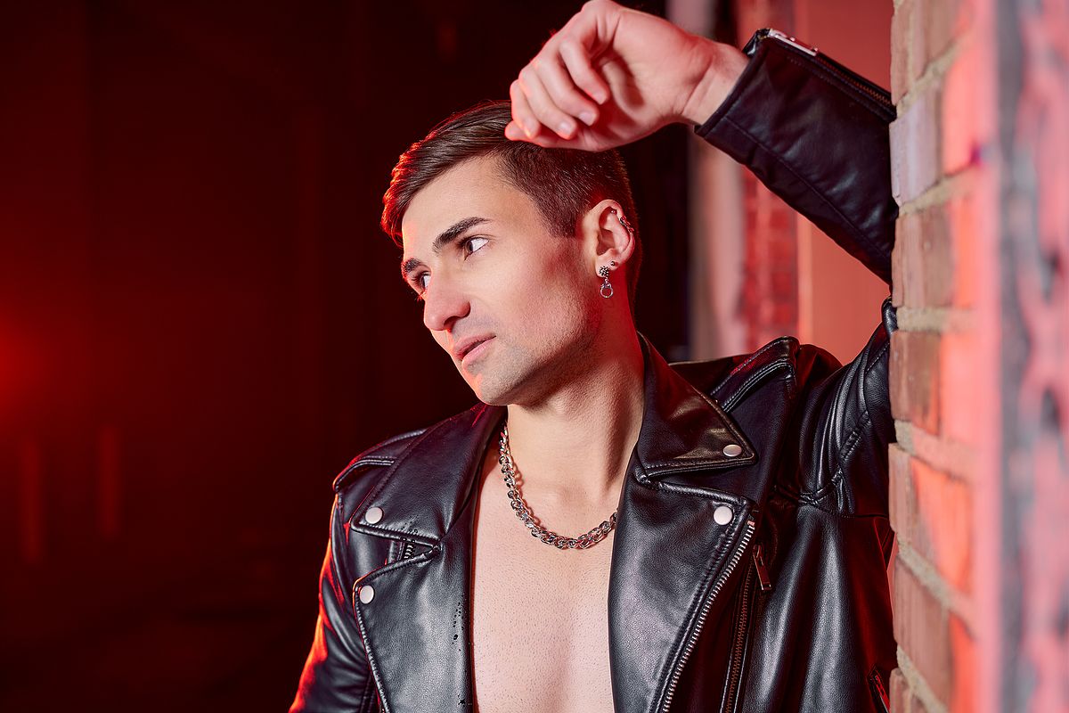 Male gay recording artist photographed outdoors at night shirtless wearing a leather jacket and leaning against a brick wall