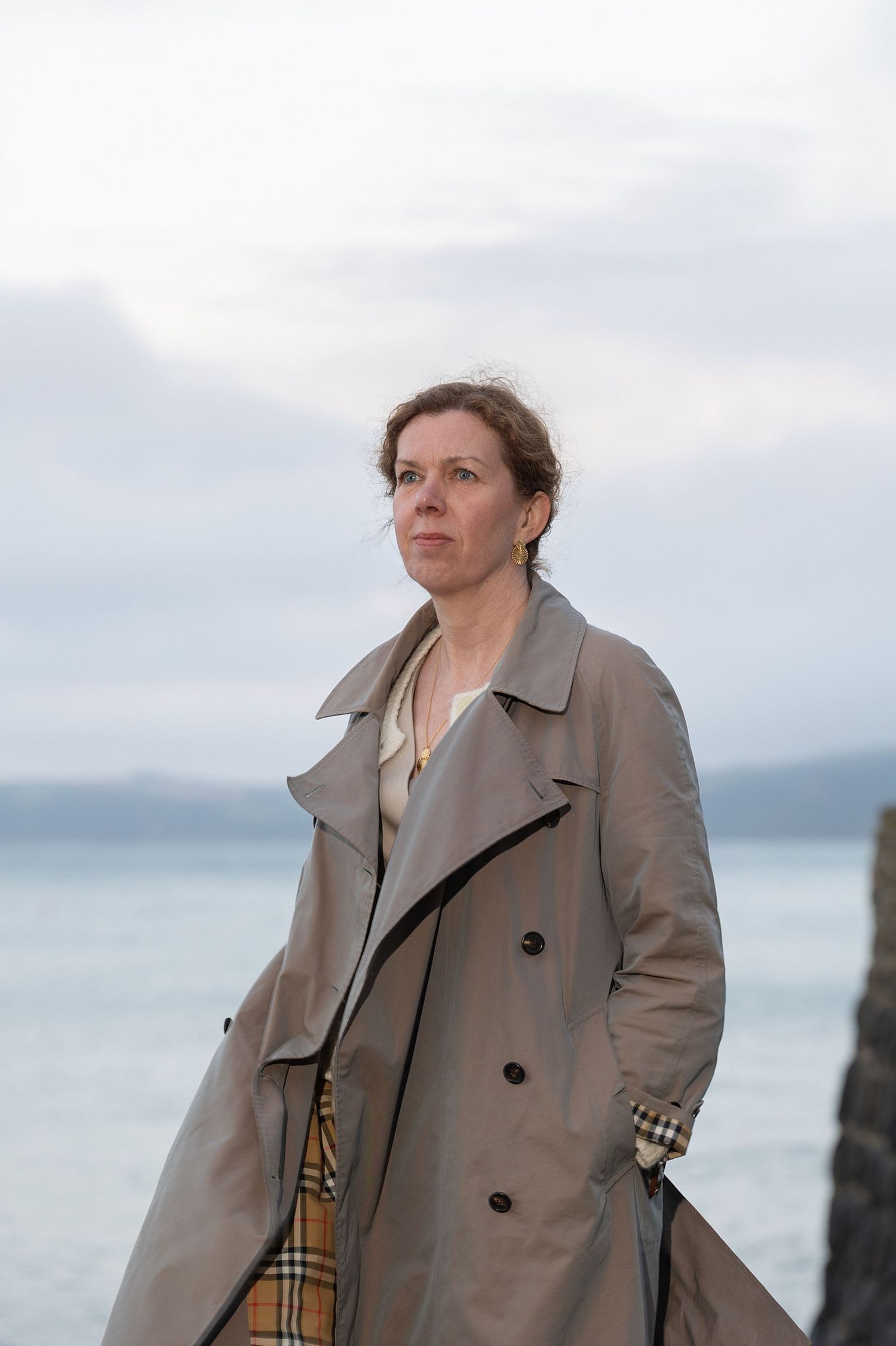 Kvinna i trenchcoat vid havet med ljus himmel, klassiskt redaktionellt portr&auml;tt med tidl&ouml;s k&auml;nsla.