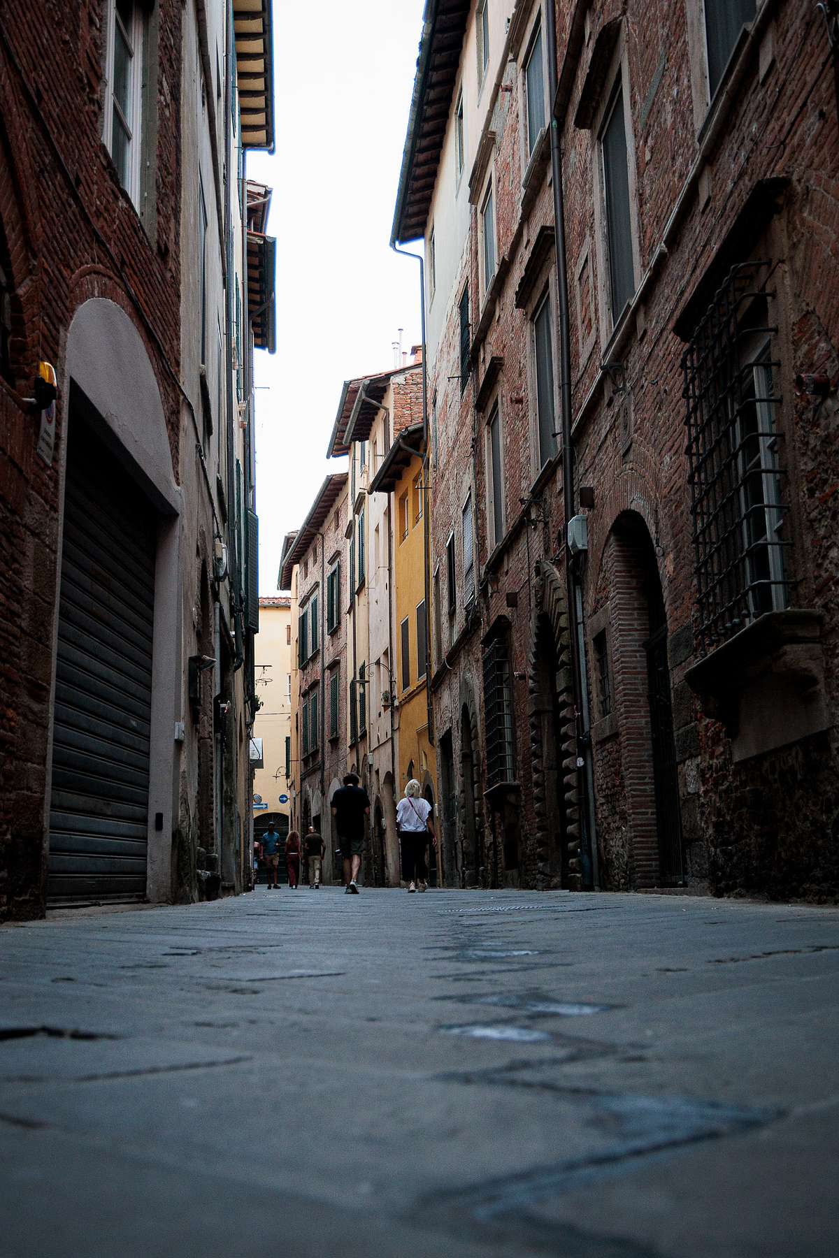 Low-angle view of a narrow cobblestone street in Lucca, Italy, lined with tall old brick and stone buildings in warm tones, including a yellow facade, with a few people walking in the distance and soft evening light creating a moody, historic atmosphere.