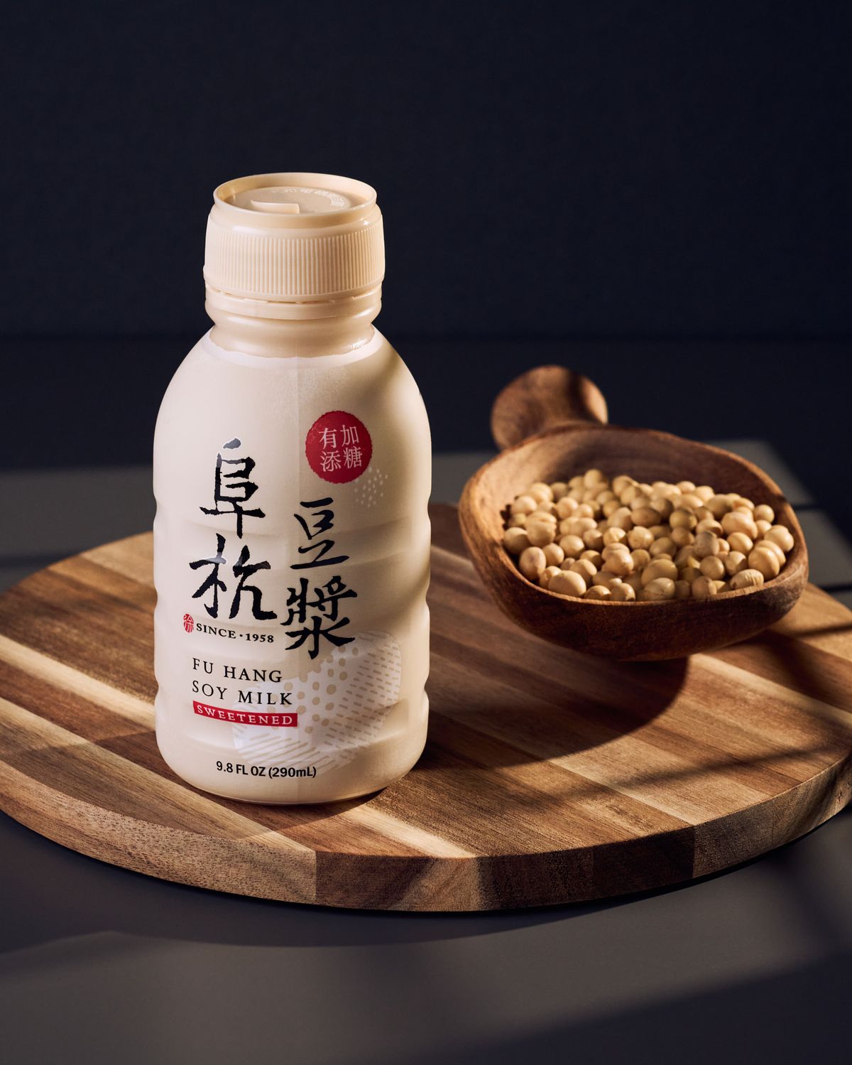 A tan bottle of soy milk and a wooden scoop of soybeans, both sitting on a wooded dish under window light.