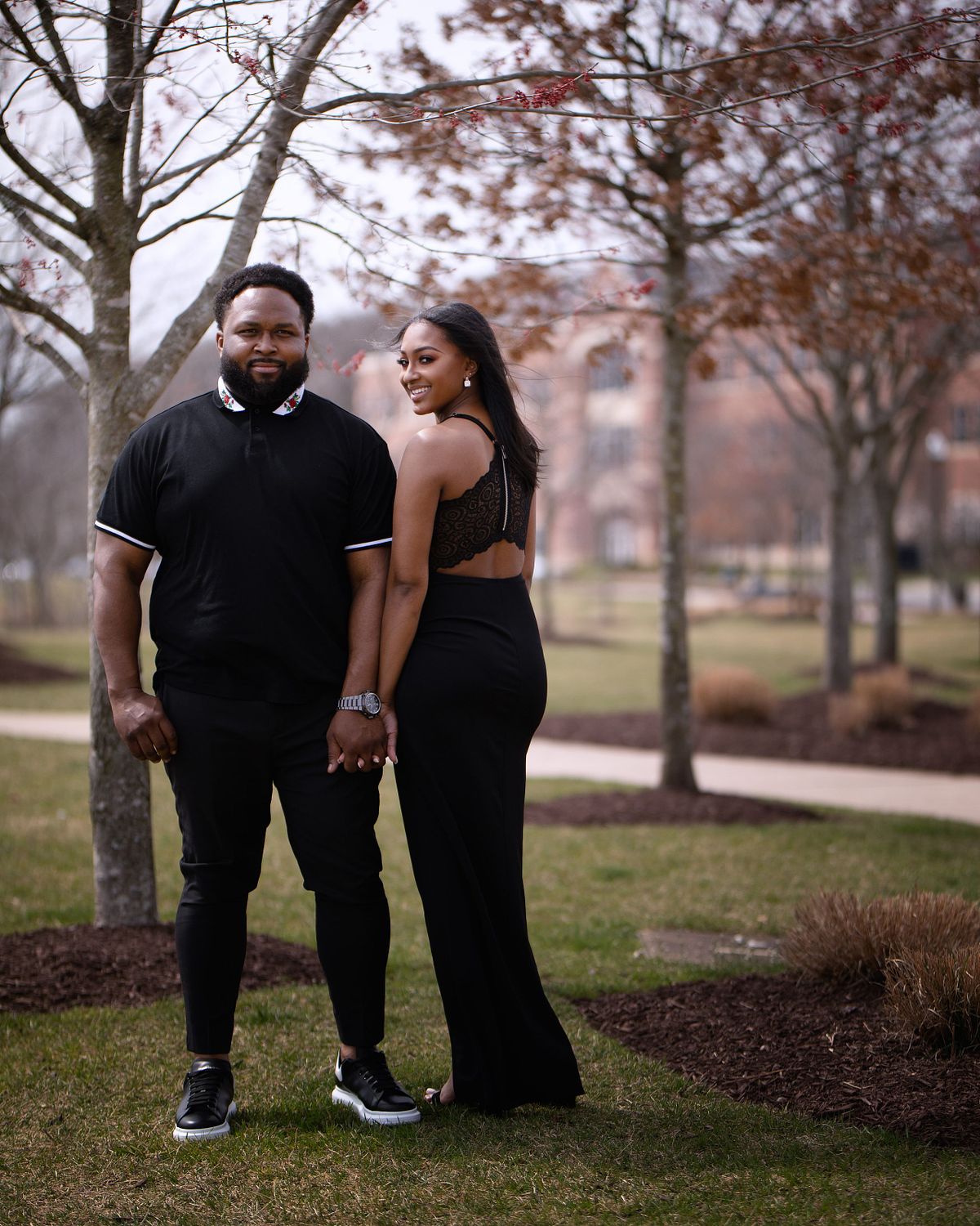 black couple during engagement session, the man is facing the camera, the couple is holding hands, both are wearing black, the woman's back is turned towards the camera but her head is turned and she is smiling
