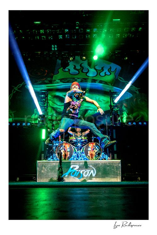 Vertical color image of Bret Michaels leaping in midair during a Poison concert with Rikki Rockett at the drum kit under vivid green stage light