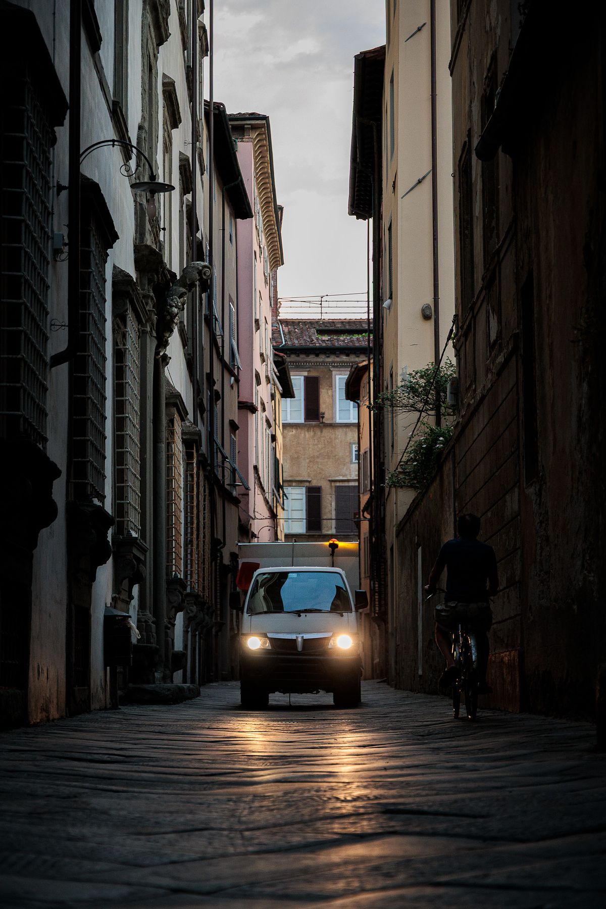 Twilight street scene in a narrow alley in Lucca, Italy, with a white van in the center of the cobblestone lane, headlights glowing warmly on the ground, a cyclist passing on the right in dark clothing, and tall beige, pink and brown buildings on both sides under an overcast evening sky, creating a calm, moody atmosphere.