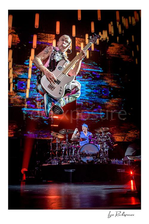 Vertical color image of Flea of Red Hot Chili Peppers leaping high with his bass above the stage, with Chad Smith visible at the drum kit below against a vivid LED backdrop