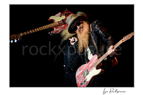 Horizontal color image of Billy Gibbons and Dusty Hill of ZZ Top performing live on stage, with Gibbons leaning into a red Telecaster while Hill lifts his red bass behind him against a dark background