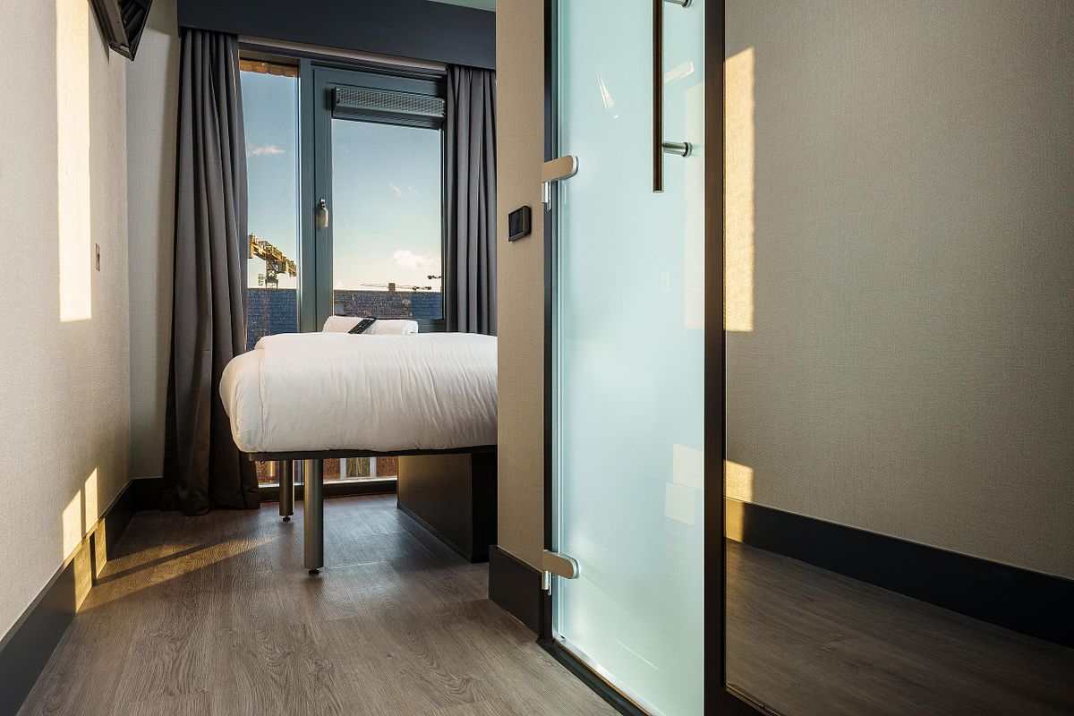 Interior view of a single room at the Easy Hotel Dublin, highlighting the natural light from a large window. A bed with white linens is partially visible. A frosted glass door to the en-suite bathroom is prominent in the foreground.