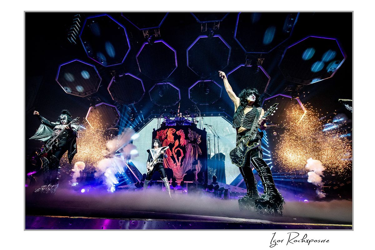 Horizontal color photograph of Gene Simmons, Paul Stanley, and Tommy Thayer performing onstage in full makeup and costumes with hexagonal lighting panels, red emblem backdrop, smoke, and pyrotechnic effects
