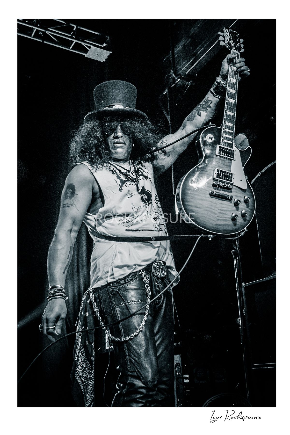Vertical concert photography of Slash holding his electric guitar high in the air with one hand in black and white concert photography under stage lighting