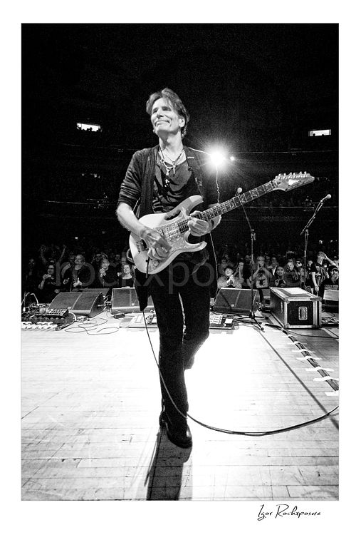 Vertical black and white image of Steve Vai walking across the stage with guitar toward the crowd and a bright backlight