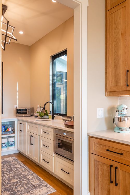 View of Pantry from kitchen showing the natural white oak cabinets transitioning into the cream colored cabinets in the pantry through the cased opening