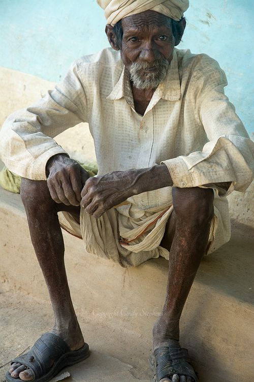 An older man in sandals and a turban sits on a low wall against the bluewashed home in Village Rancha, India, gazing pointedly at the camera.