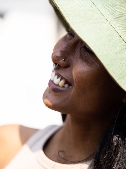 A headshot of an Indian woman smiling, with a nose ring, wearing a green bucket hat on a sunny day.