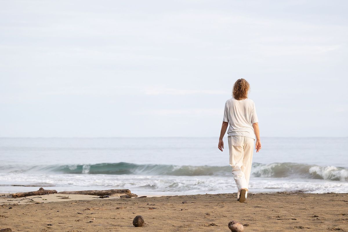 Redaktionellt milj&ouml;portr&auml;tt bakifr&aring;n: person g&aring;r mot havet p&aring; strand i mjukt ljus, minimalistisk komposition.