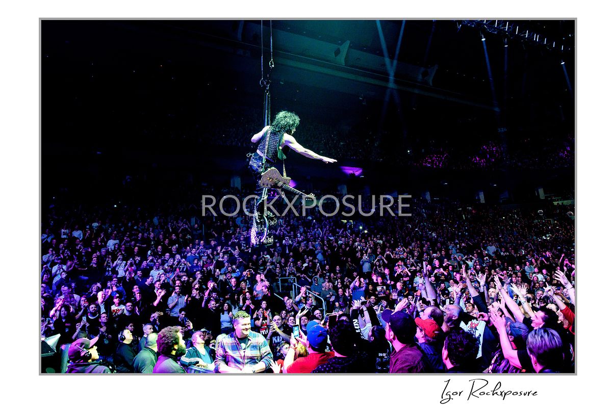 Dramatic horizontal concert photography of Paul Stanley from KISS flying above a packed arena crowd while playing guitar suspended by a cable with blue stage lighting illuminating the audience below