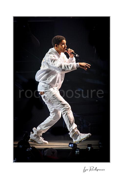 Vertical color image of Drake performing live on stage in white outfit holding microphone mid-stride with dynamic movement