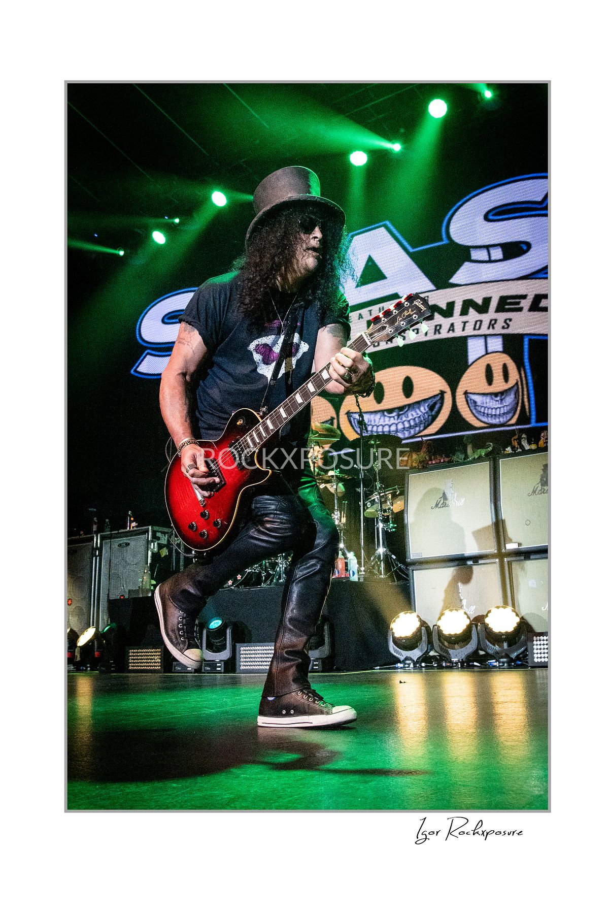 Vertical concert photography of Slash playing electric guitar in a wide stance under green stage lighting with Slash stage backdrop