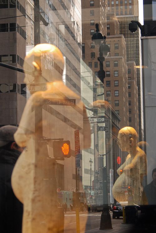 Artistic humanoid shapes juxtaposed against classic NYC skyscrapers and a busy street reflected in the background
