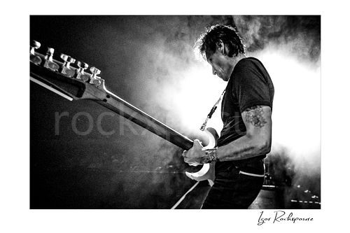 Horizontal black and white image of Steve Vai playing guitar in side profile through stage smoke with strong backlight