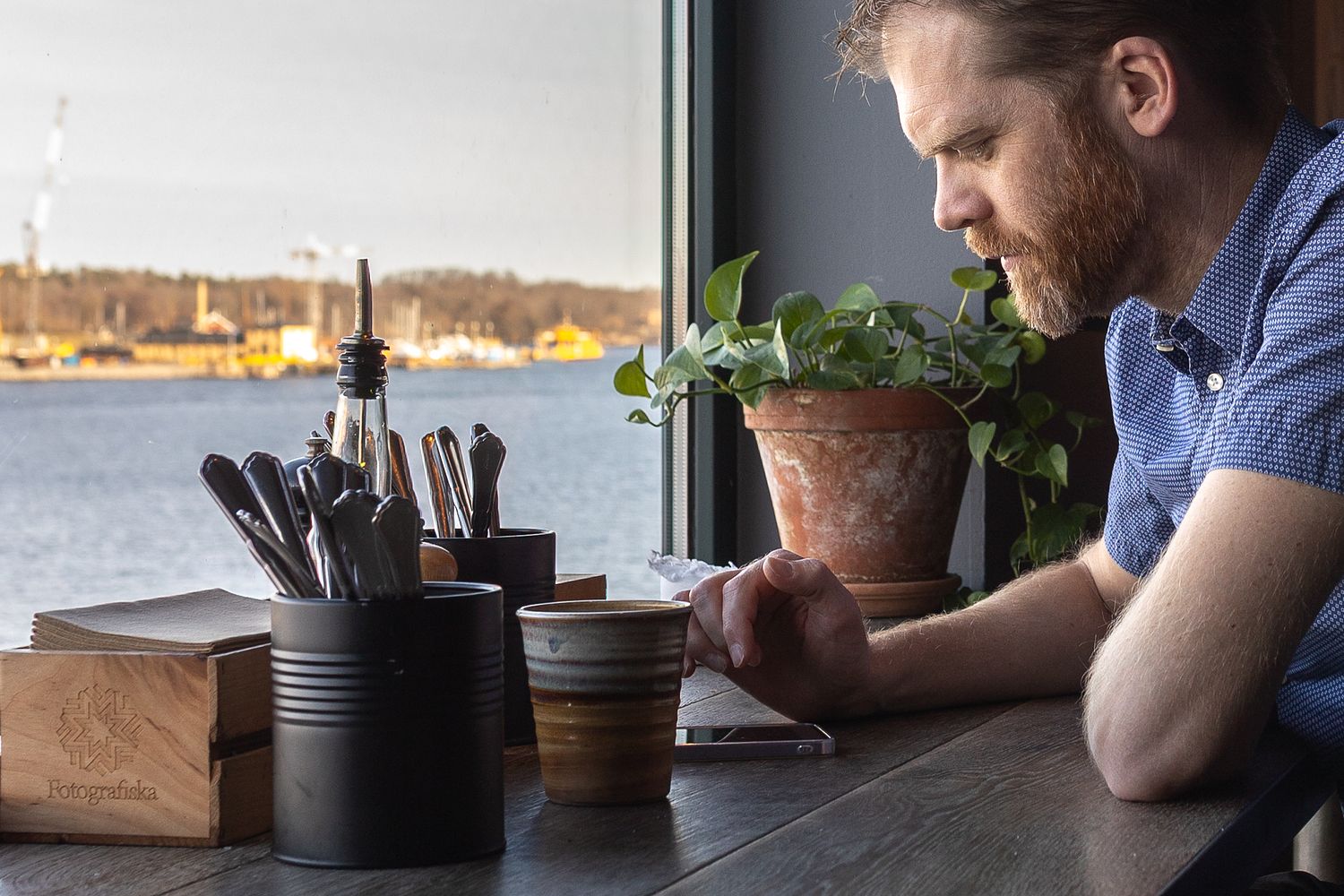 Lifestyle portrait of a man sitting at a café table by a window overlooking water, photographed in Stockholm by Mats Karlsson.
