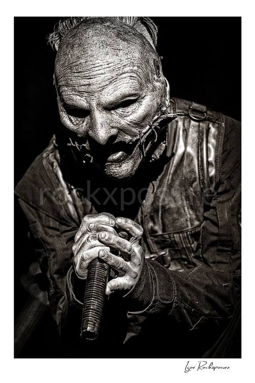 Vertical black and white image of Corey Taylor of Slipknot in a tight masked close-up gripping a handheld microphone with both hands