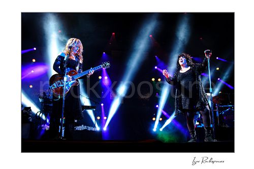 Horizontal color image of Nancy Wilson and Ann Wilson performing live on stage during a Heart concert with electric guitar, microphone and vivid blue and purple stage lights
