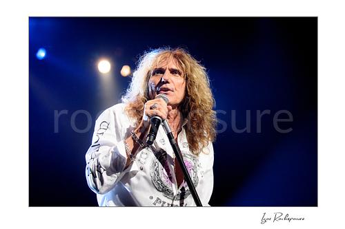 Horizontal color close-up image of David Coverdale of Whitesnake performing live on stage, singing into a microphone while holding a mic stand under blue concert lighting