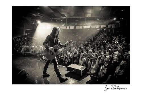 Horizontal black and white image of Tom Keifer performing live on stage with an acoustic guitar, walking toward the crowd under a bright spotlight with fans reaching forward
