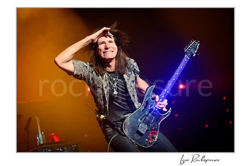 Horizontal color image of Steve Vai playing guitar with windswept hair and one hand raised to his forehead under warm stage lighting