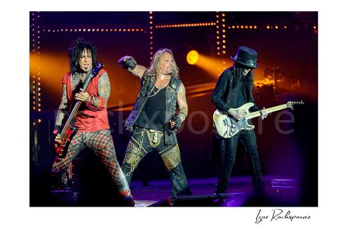 Horizontal color image of Nikki Sixx, Vince Neil and Mick Mars performing live on stage during a Mötley Crüe concert with bass guitar, microphone and warm concert lighting