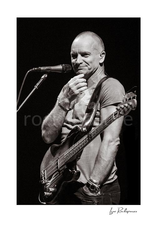 Vertical black and white image of Sting holding a bass guitar beside a microphone in a quiet live performance portrait