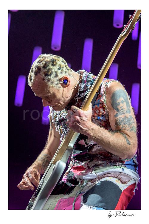 Vertical color image of Flea of Red Hot Chili Peppers playing bass in a tight live performance close-up, with shaved patterned hair, vivid stage light, and glowing LED tubes behind him