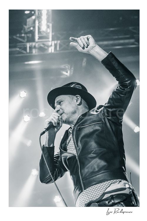 Vertical black and white image of Gord Downie of The Tragically Hip singing into a microphone with one arm raised, wearing a hat and leather jacket under bright stage lights