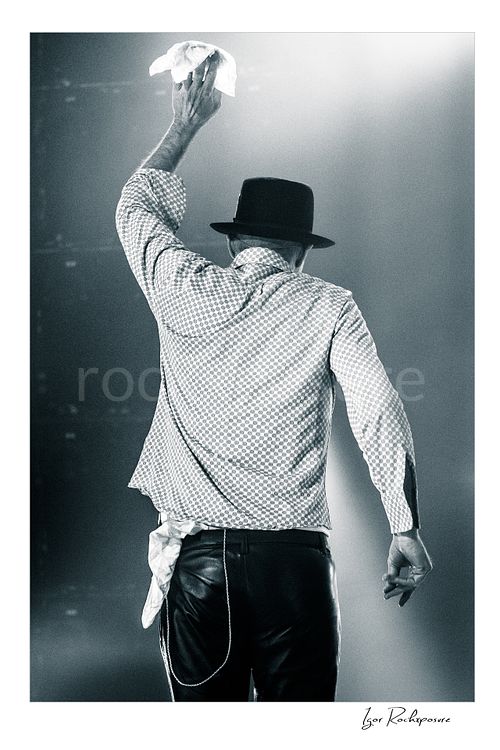 Vertical black and white image of Gord Downie of The Tragically Hip seen from behind on stage, raising a white cloth in a final farewell gesture under soft concert light