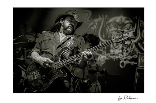 Horizontal black and white image of Lemmy Kilmister of Motörhead performing live on stage, wearing a cowboy hat and playing bass guitar with the band backdrop visible behind him