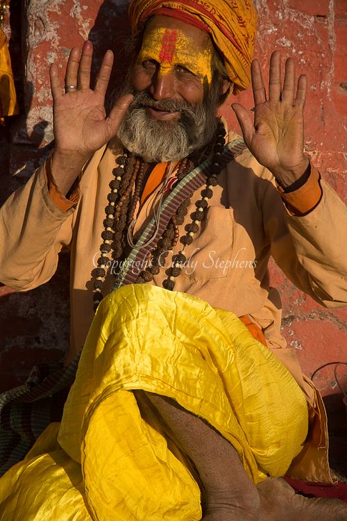 A sadhu in traditional costume with a painted face is bathed in sunset light, raising his hands near the banks of the Bagmati River at Pashupatinath Temple in Nepal.