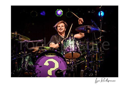 Horizontal color image of Rikki Rockett performing at the drum kit during a Poison concert, raising a drumstick under bright stage lights