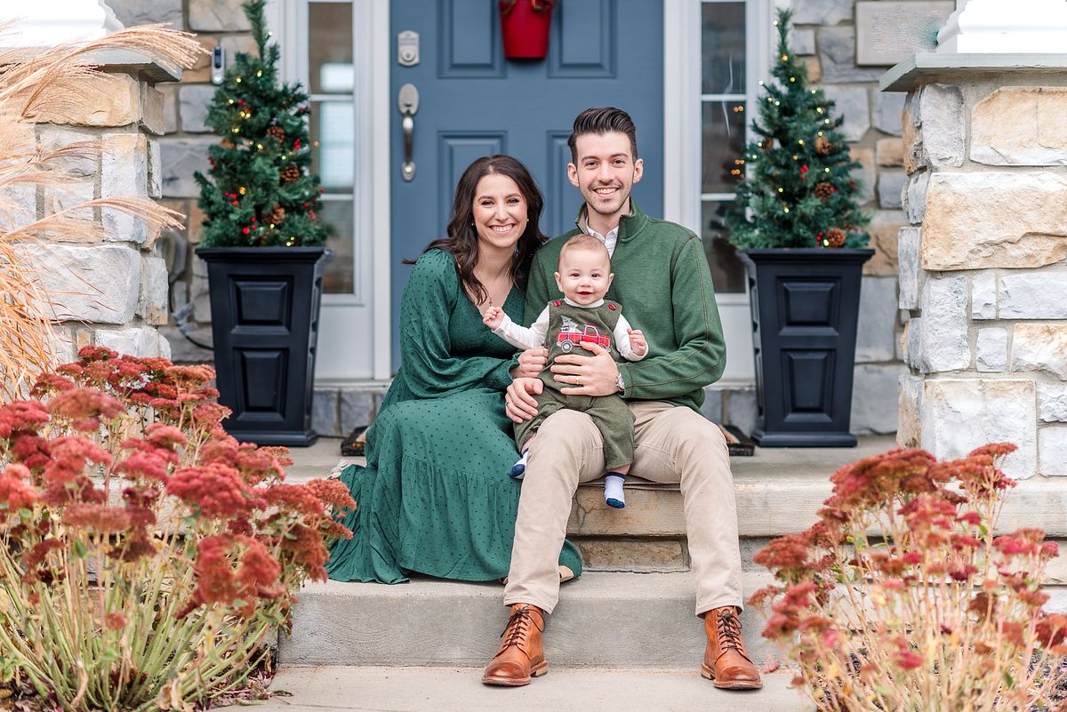 Husband and wife sitting on porch steps with their baby and Christmas decor with Cranberry Township, PA newborn photographer