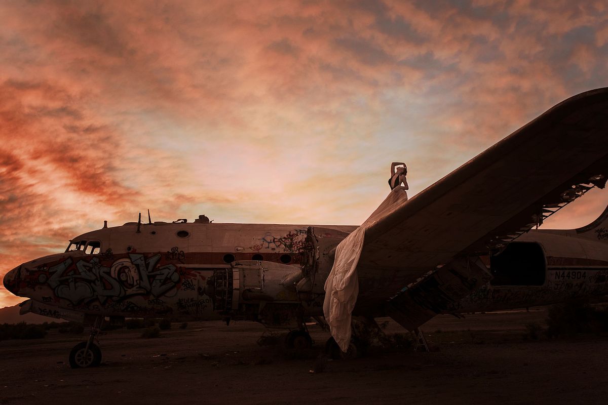 Boudoir photo of a woman sitting confidently on the wing of an abandoned airplane at sunset, radiating strength and beauty in the Arizona desert. Wearing a parachute skirt.