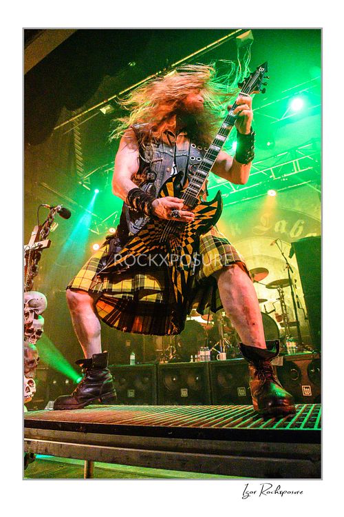 Low-angle vertical shot of Zakk Wylde standing with legs wide apart on a grated stage riser, wearing a kilt and playing guitar with his hair flying back under strong green lighting.