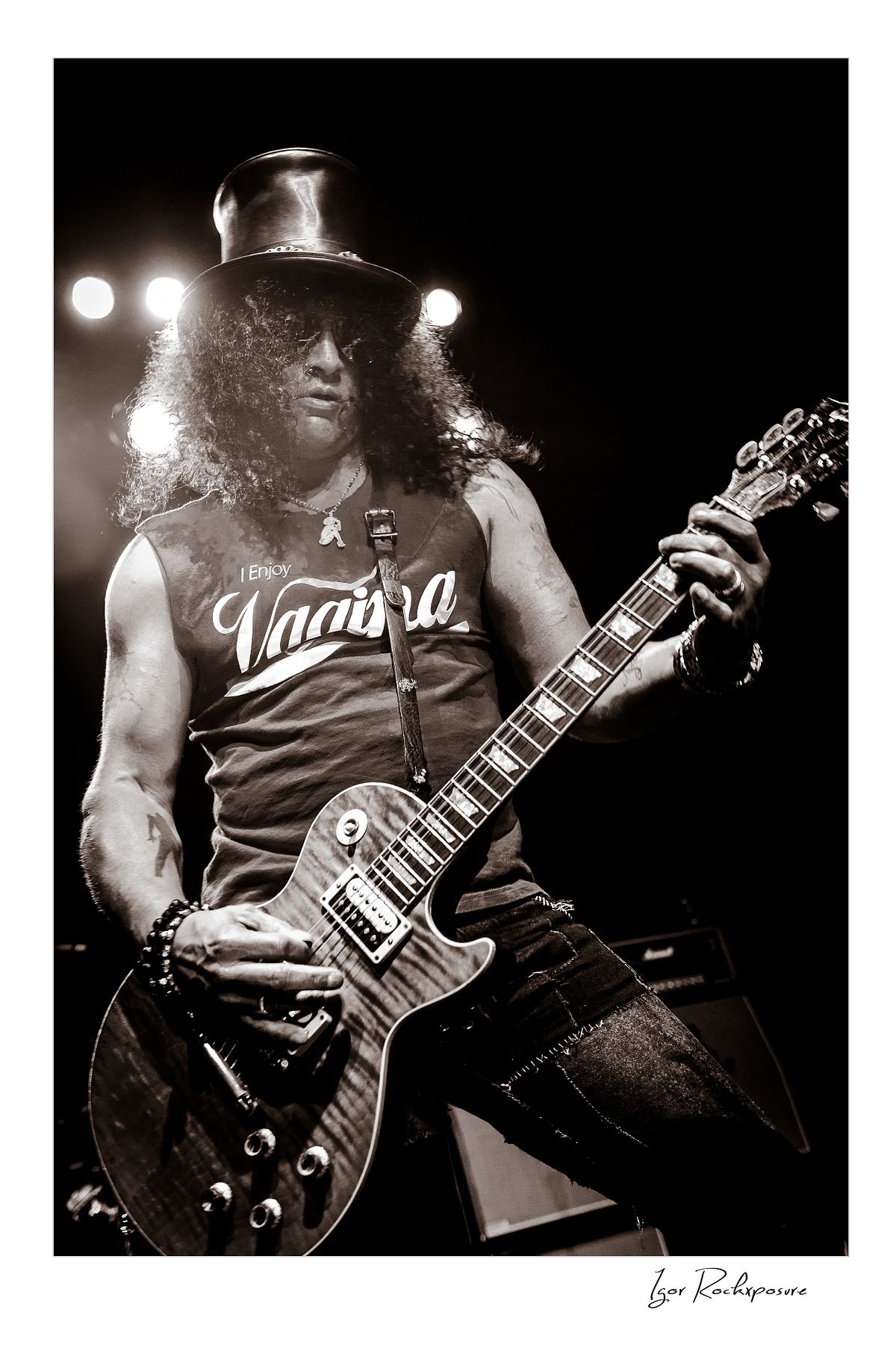 Vertical concert photography of Slash playing electric guitar in black and white photography wearing his signature top hat under dramatic stage lighting