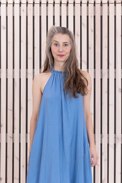 Natural light environmental portrait of a woman in a blue dress standing against a modern wooden slat wall, photographed in Stockholm by Mats Karlsson.