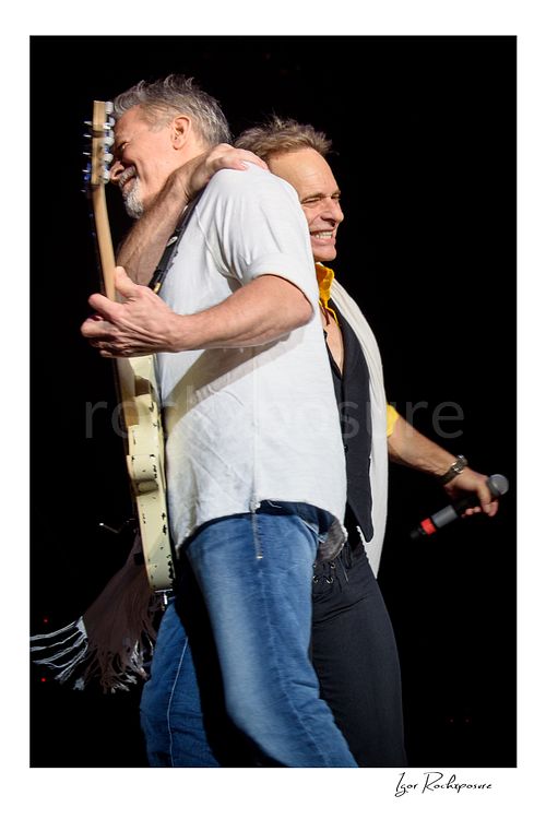 Vertical color image of Eddie Van Halen and David Lee Roth of Van Halen sharing an onstage embrace with Eddie holding a white electric guitar