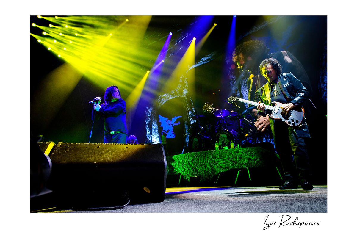 Tony Iommi playing guitar on stage while Ozzy Osbourne sings beside him under dramatic yellow and blue concert lighting during a Black Sabbath performance