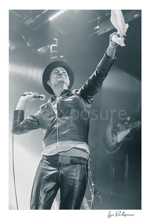 Vertical black and white image of Gord Downie of The Tragically Hip holding a microphone and lifting a white cloth high on stage, wearing a hat and leather jacket in bright concert haze