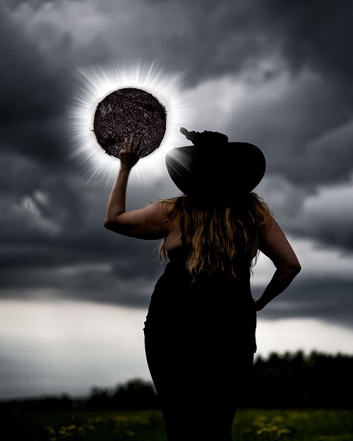 A witch reaching up to a black sun with a bright white halo.